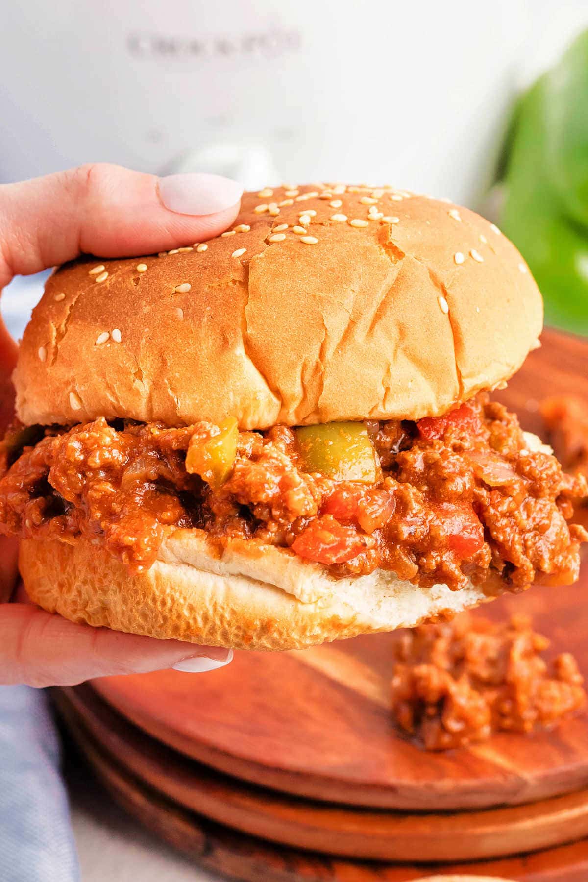 A hand holding a messy Sloppy Joe sandwich on a sesame seed bun.