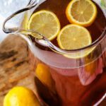 A pitcher of lemon iced tea with lemon slices floating on top on a table with fresh lemons and glasses filled with ice cubes.