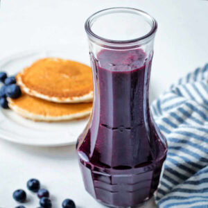 Blueberry syrup in a carafe on a table with a plate of pancakes.