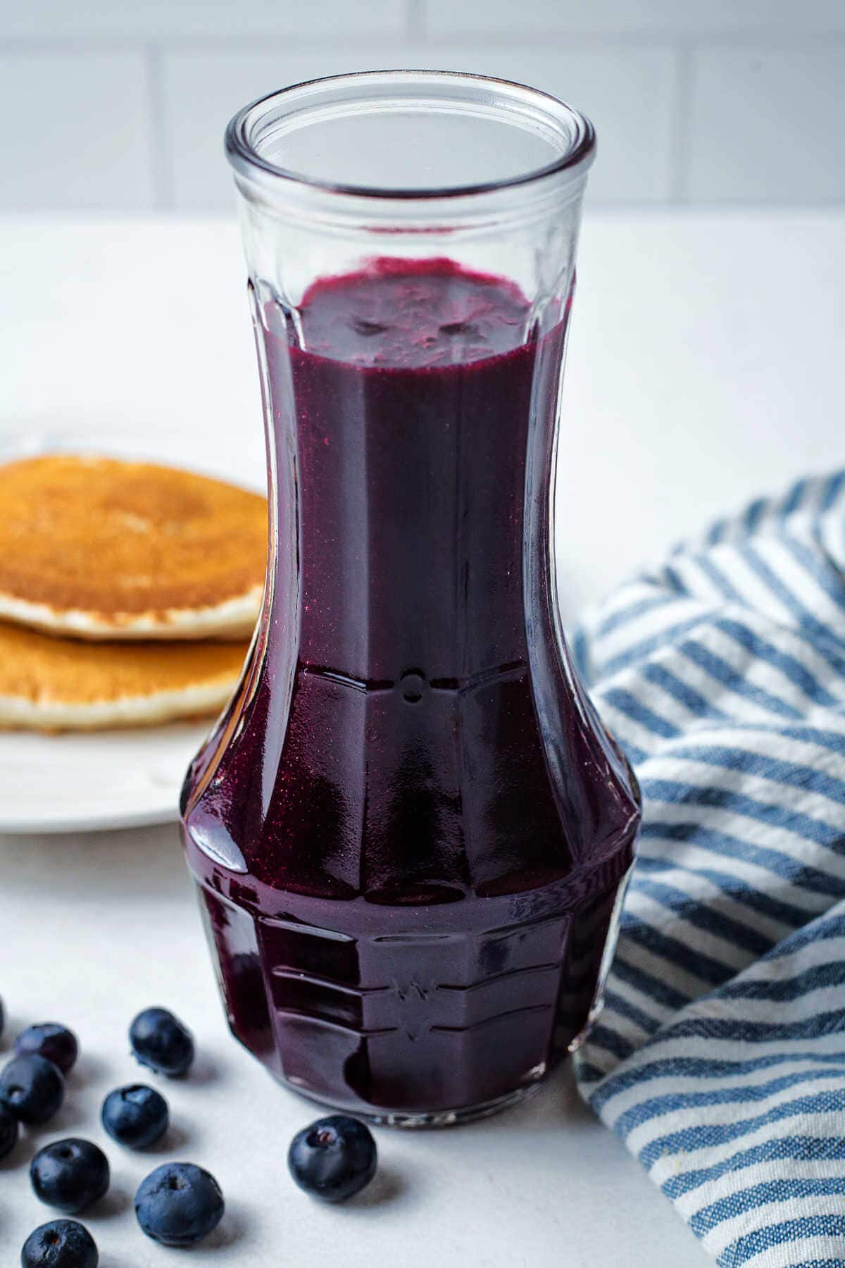 Blueberry syrup in a carafe on a table with a plate of pancakes.