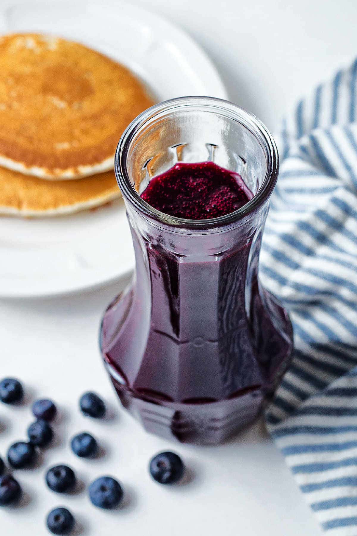 Blueberry syrup in a carafe on a table with a plate of pancakes.