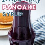 Blueberry syrup in a carafe on a table with a plate of pancakes.