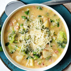 Broccoli Potato Soup garnished with shredded Parmesan cheese in a bowl sitting on a blue dinner plate.