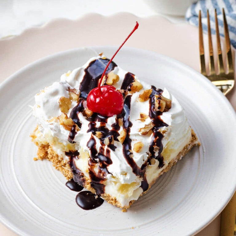 A slice of banana split cake drizzled with chocolate witih a cherry on top on a plate.
