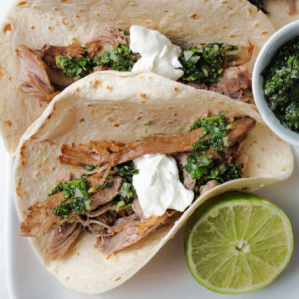 Close up of a pulled pork taco with chimichurri and sour cream on top next to a lime half.