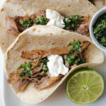 Close up of a pulled pork taco with chimichurri and sour cream on top next to a lime half.