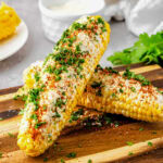 Two pieces of corn leaning on each other, each with toppings like mayo, cheese, tajin and cilantro.