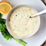 Homemade tartar sauce in a small white bowl placed on a larger white plate.
