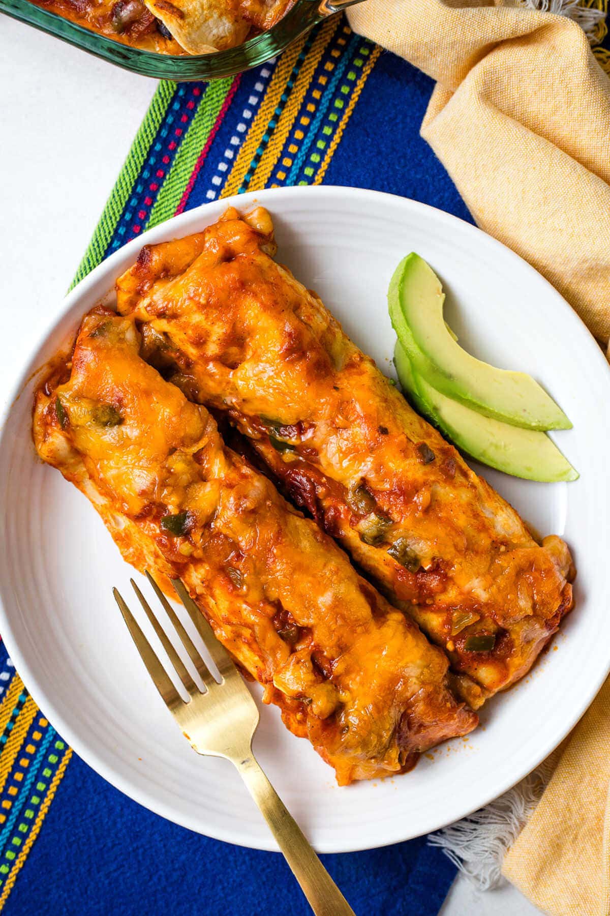 Two chicken and black bean enchiladas on a white plate served with avocado on the side.