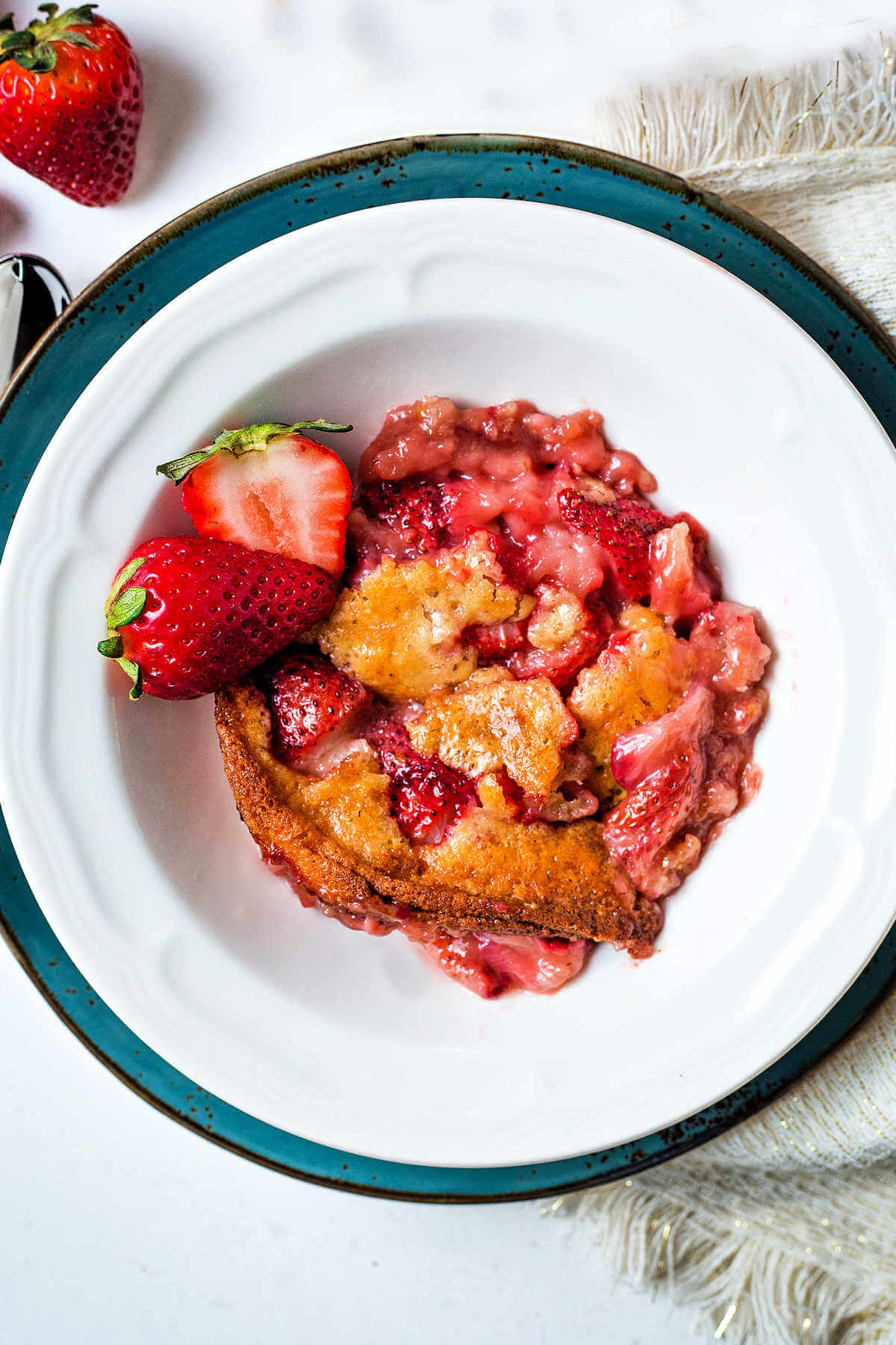 Overhead shot of strawberry cobbler in a white plate with fresh strawberries on the side.