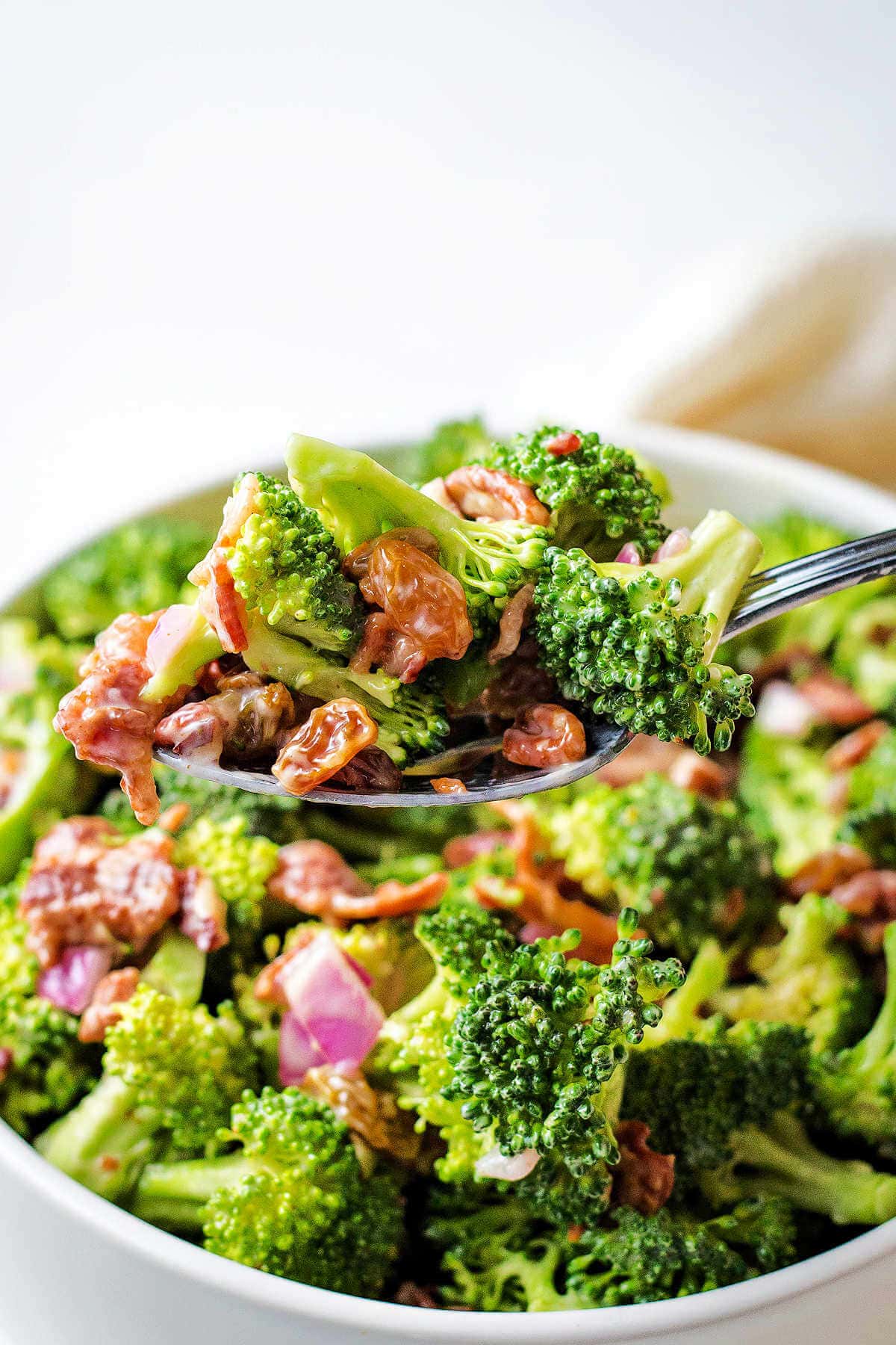 A spoon lifting broccoli salad out of a bowl on a table.