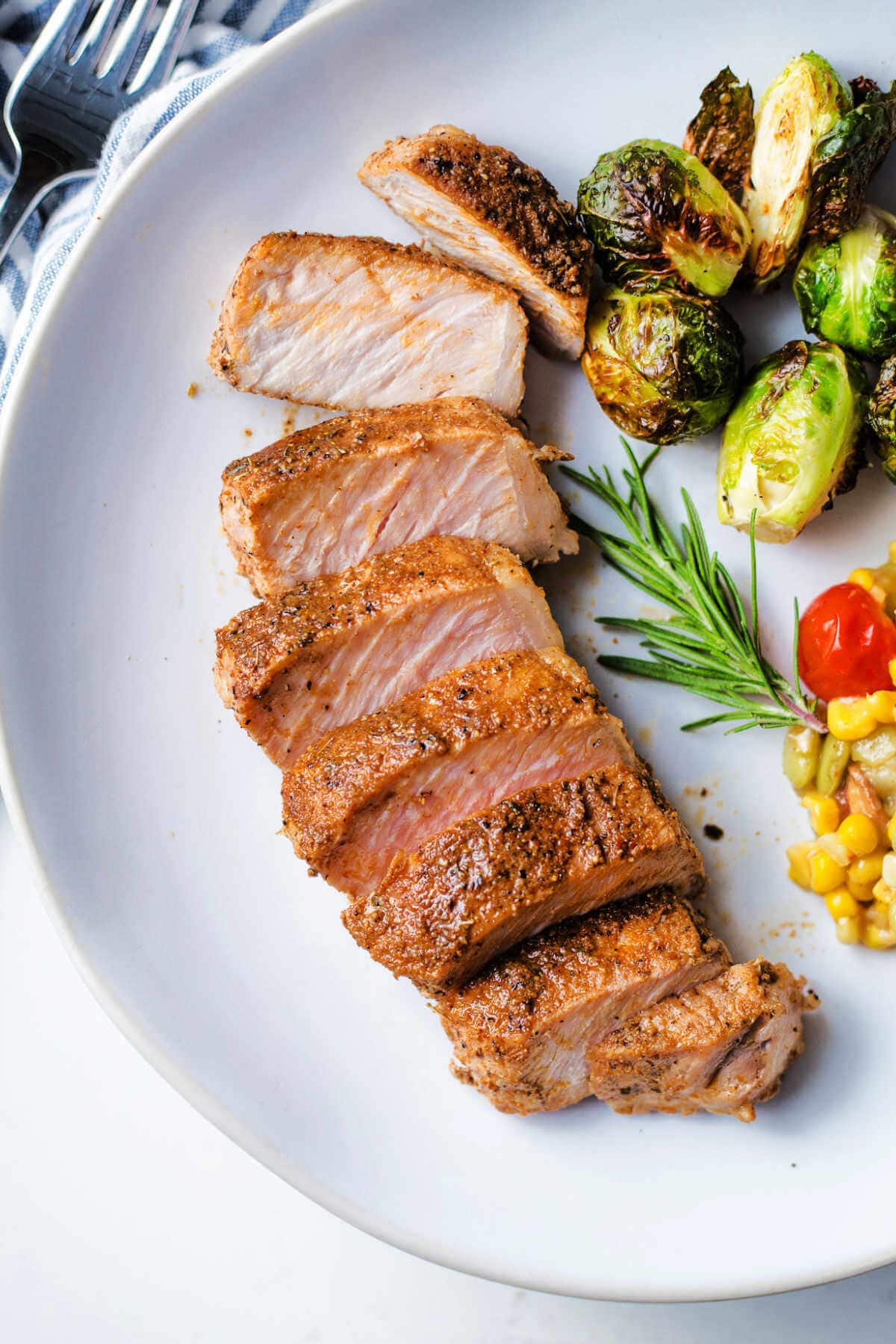 Sliced pork loin chop on a plate next to sides of Brussels sprouts and corn.