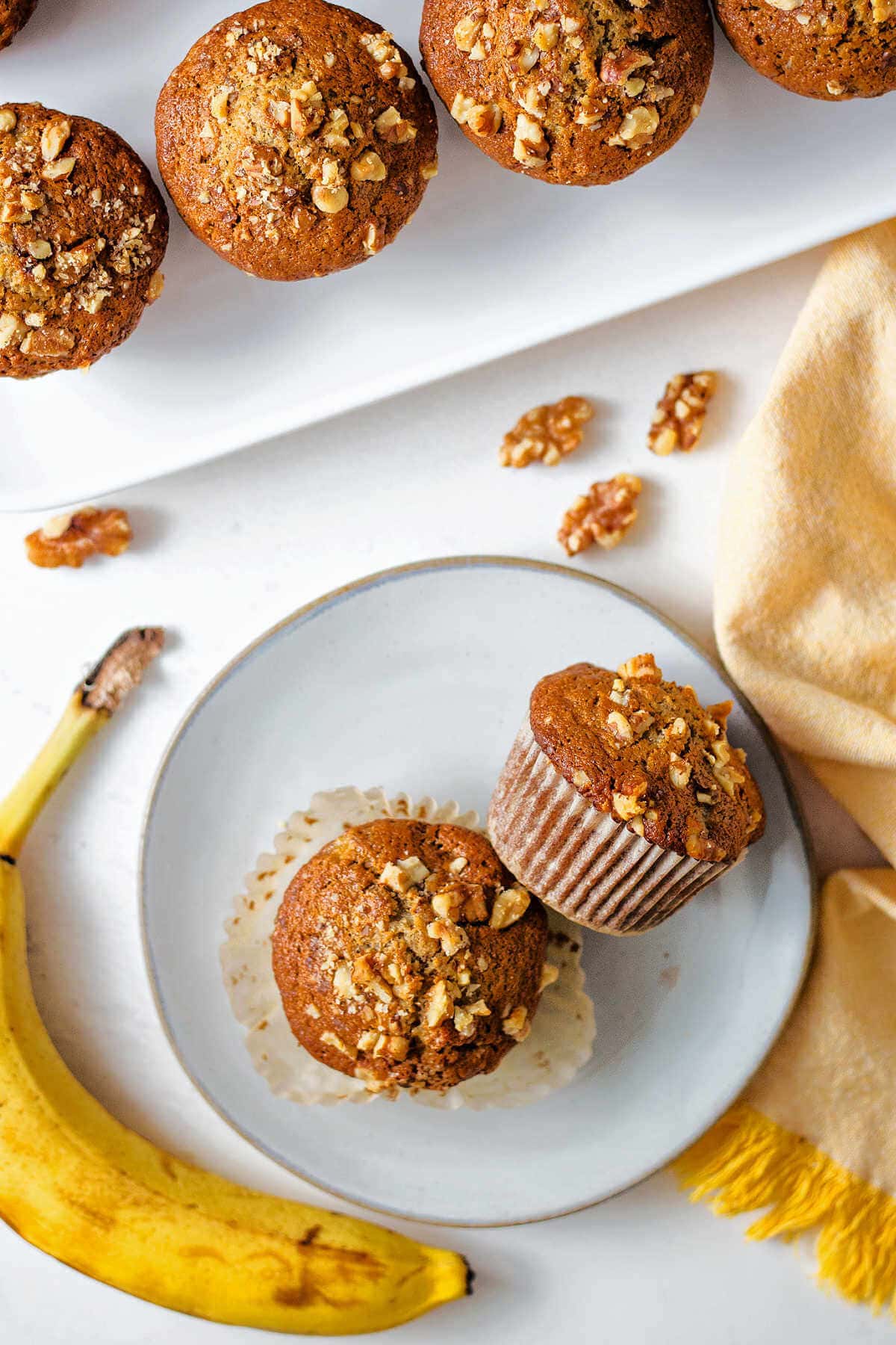 Two banana nut muffins on a plate with more on a serving platter.