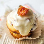 A Maple-Glazed Bacon Muffin garnished with a bacon piece on a paper cupcake liner.
