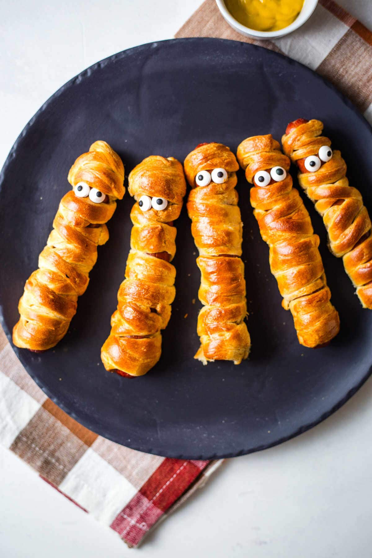 Mummy dogs with edible eyes served on a black plate.