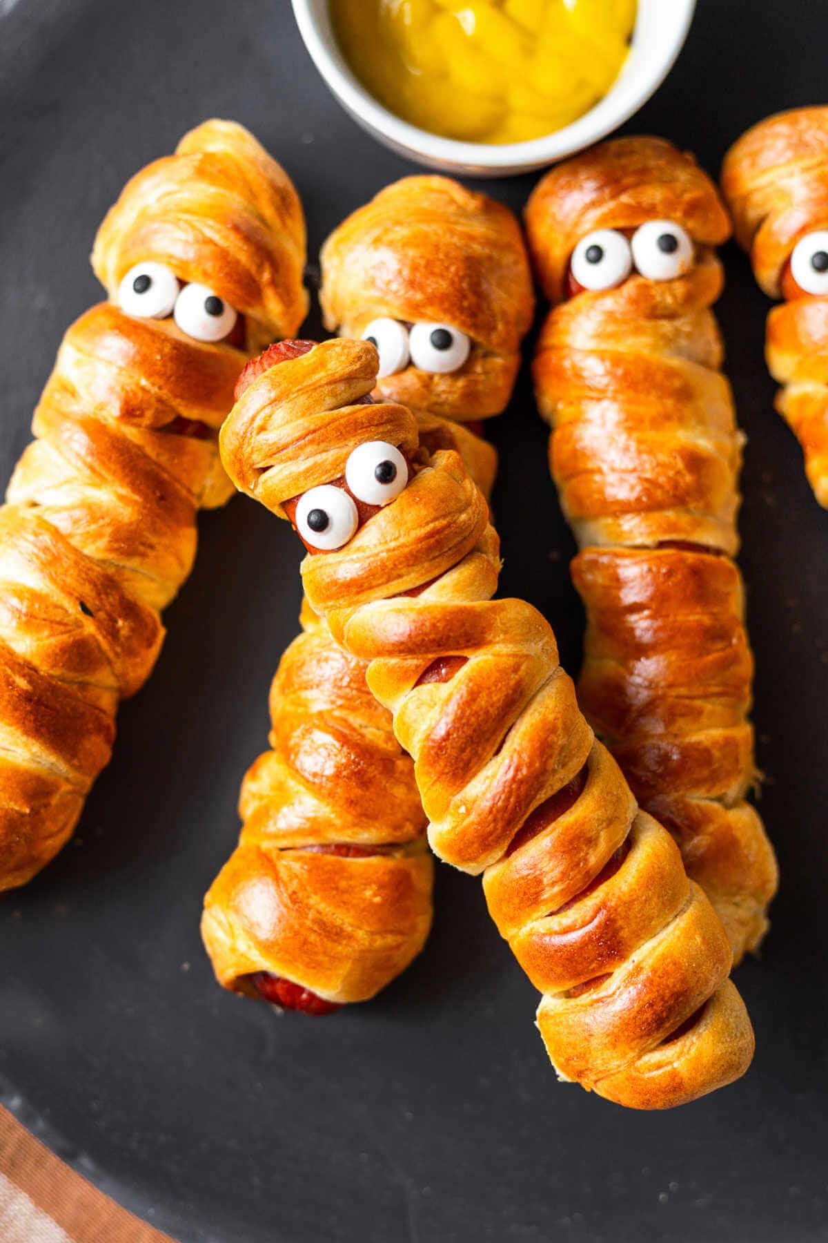 Mummy dogs served on a plate with mustard with one mummy dog leaning on the others.
