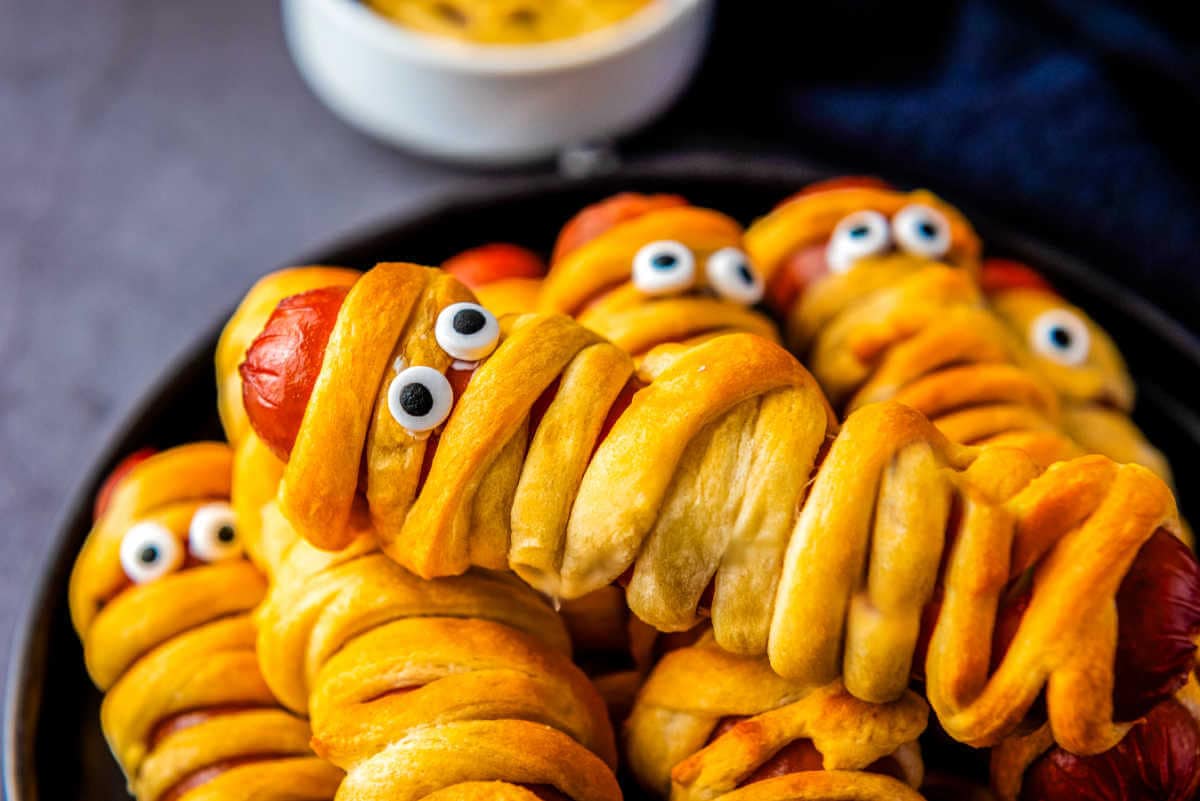 Crescent roll hot dogs with edible eyes stacked on top of each other on a plate.