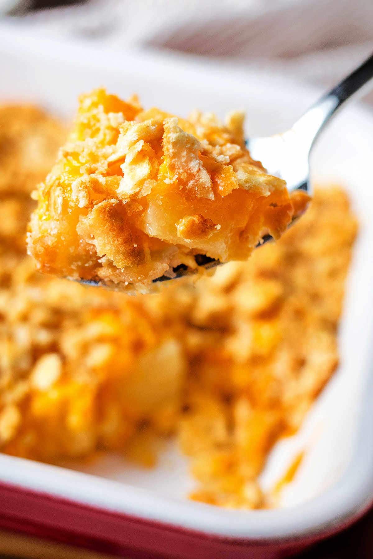 Spoon with a portion of pineapple casserole with the casserole in the background.