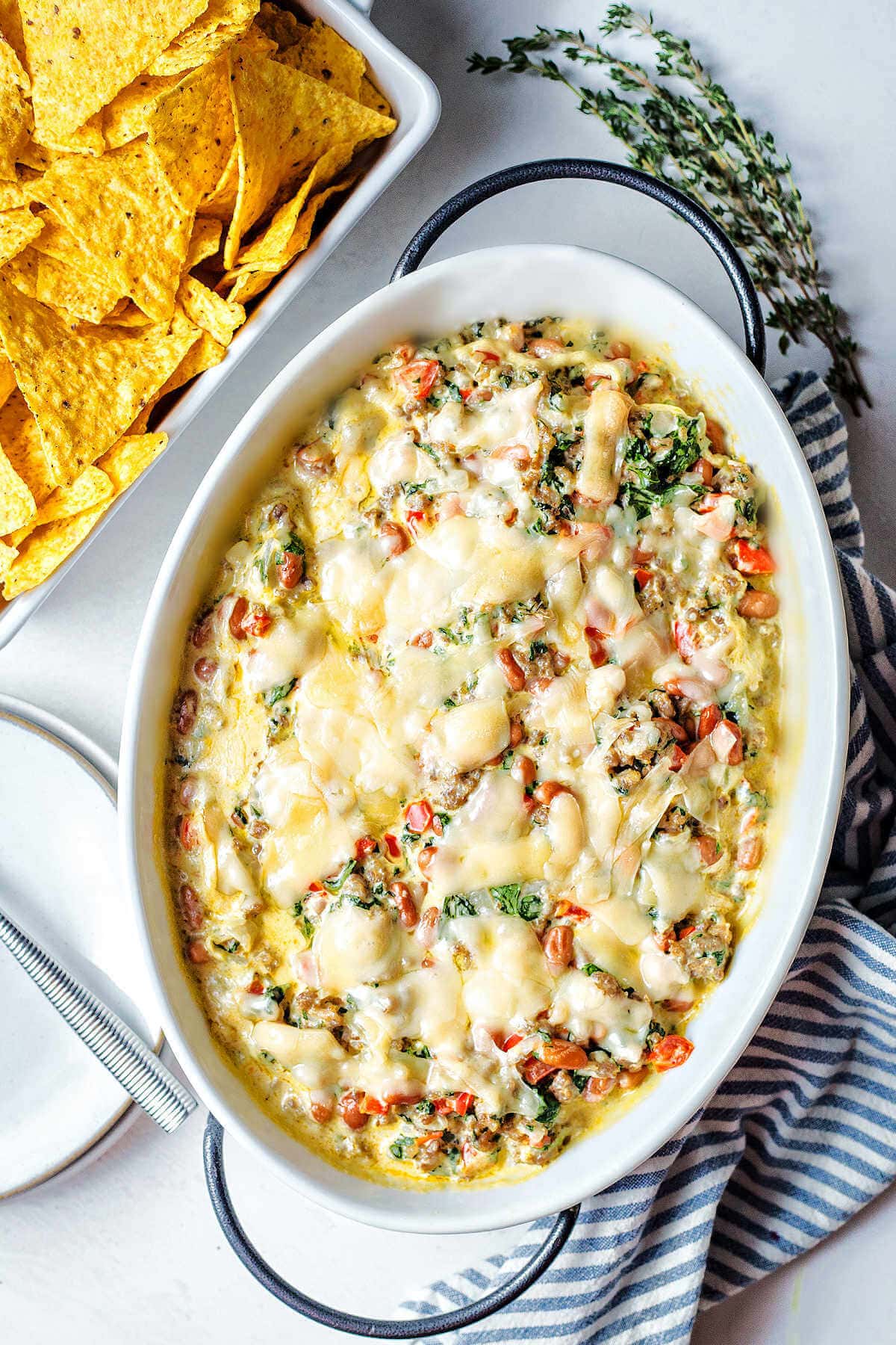 Sausage dip with beans and spinach in a baking dish next to a bowl of chips.
