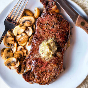 A New York strip steak cooked in cast iron on a plate with mushrooms and butter on top.
