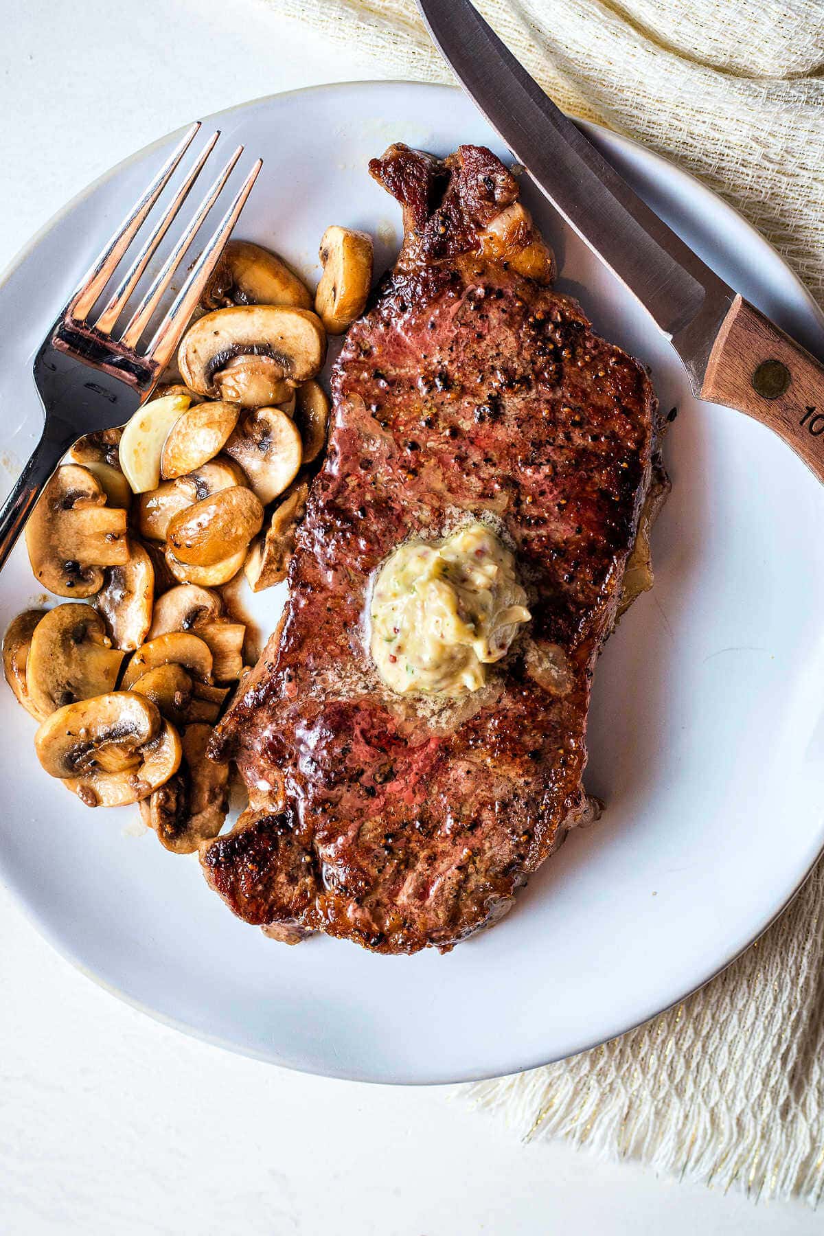 A New York strip steak cooked in cast iron on a plate with mushrooms and butter on top.