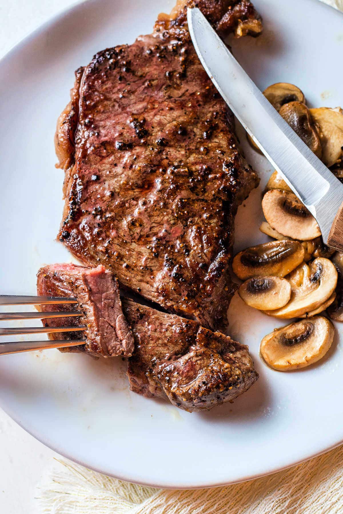 A New York strip steak cooked in cast iron on a plate with a steak knife and a piece cut and pierced on a fork.