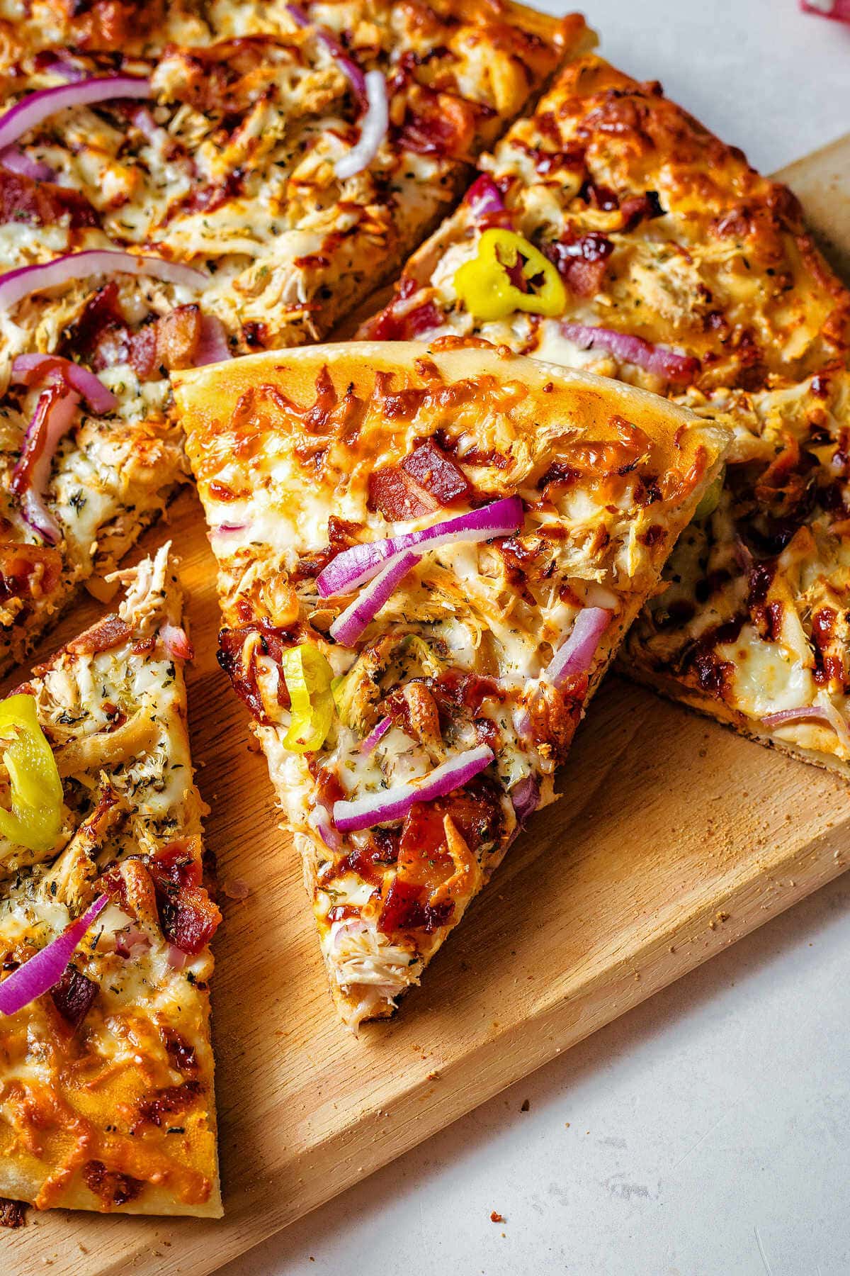 Slice of BBQ chicken pizza leaning on the other slices on a wooden board.