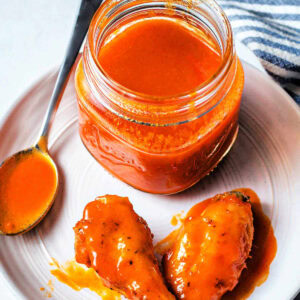 Jar of buffalo wing sauce on a plate next to two wings with buffalo sauce on them.