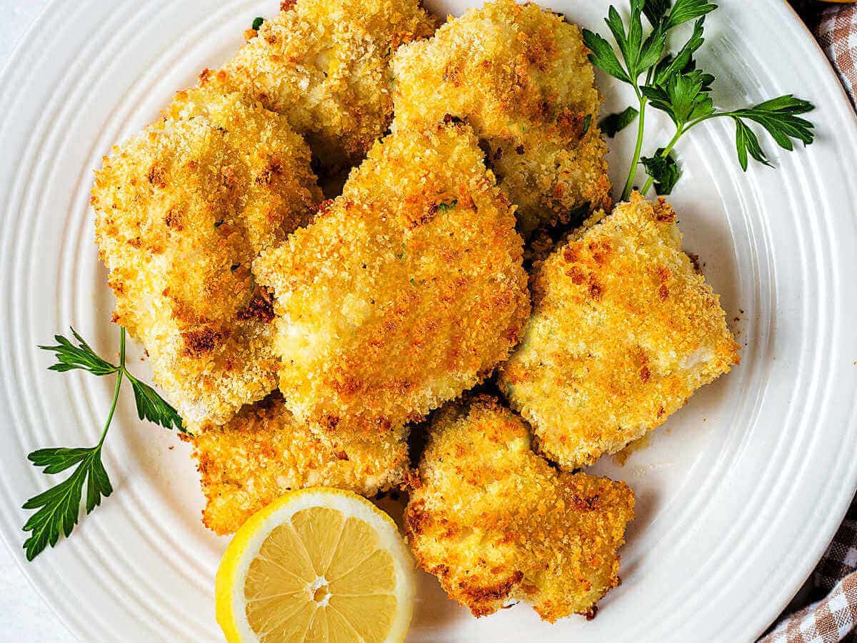 A platter of oven fried fish filets garnished with parsley and lemon halves on a table with a bowl of tartar sauce.