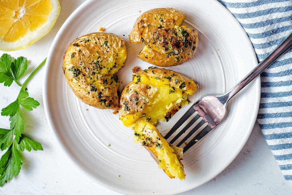 Three smashed potatoes with garlic and lemon on a plate with a fork taking a bite of one.