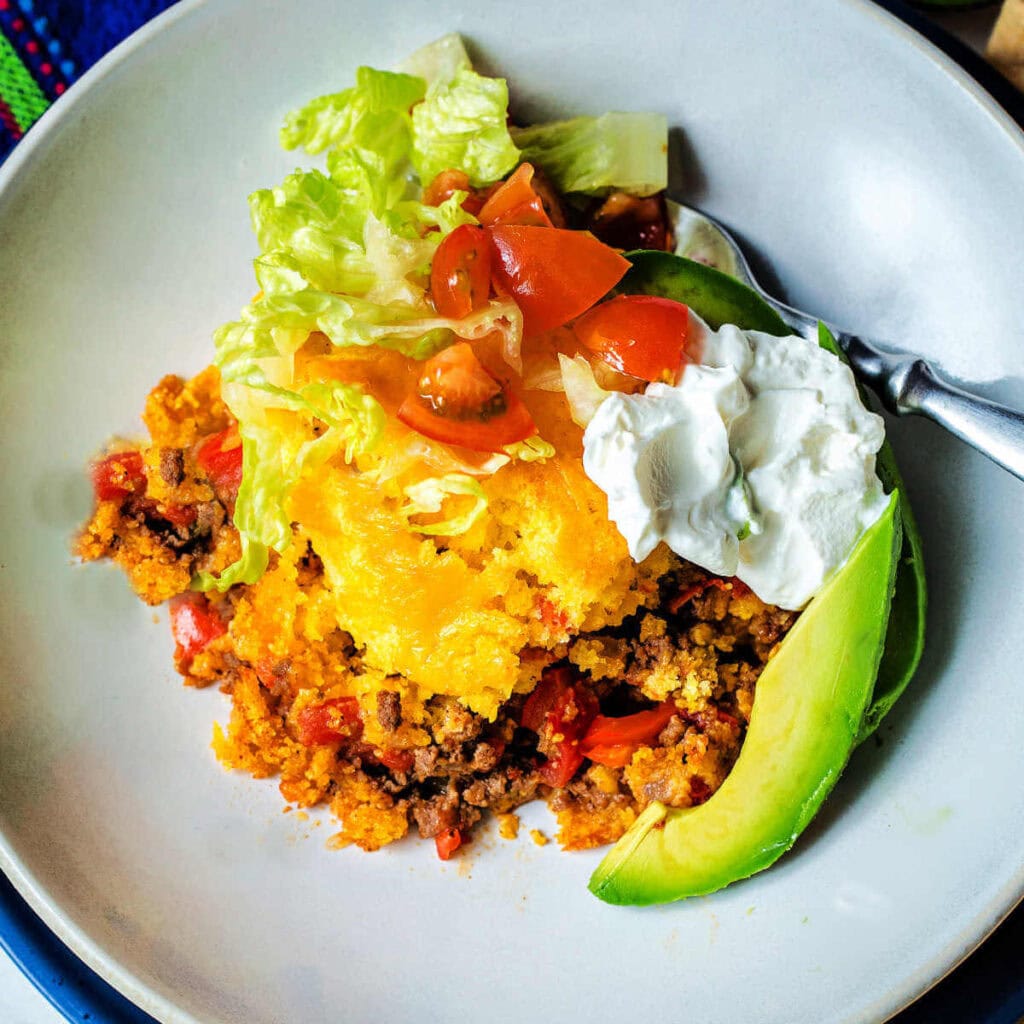 Tamale Pie serving on a plate garnished with chopped lettuce, tomatoes, avocado slices, and a dollop of sour cream.
