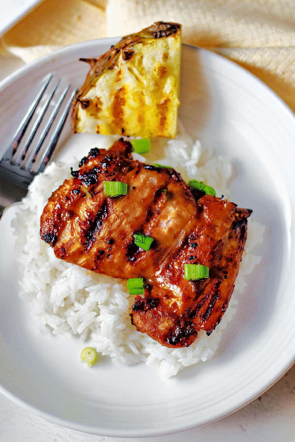Huli huli chicken breast served on rice with a side of pineapple on a plate.