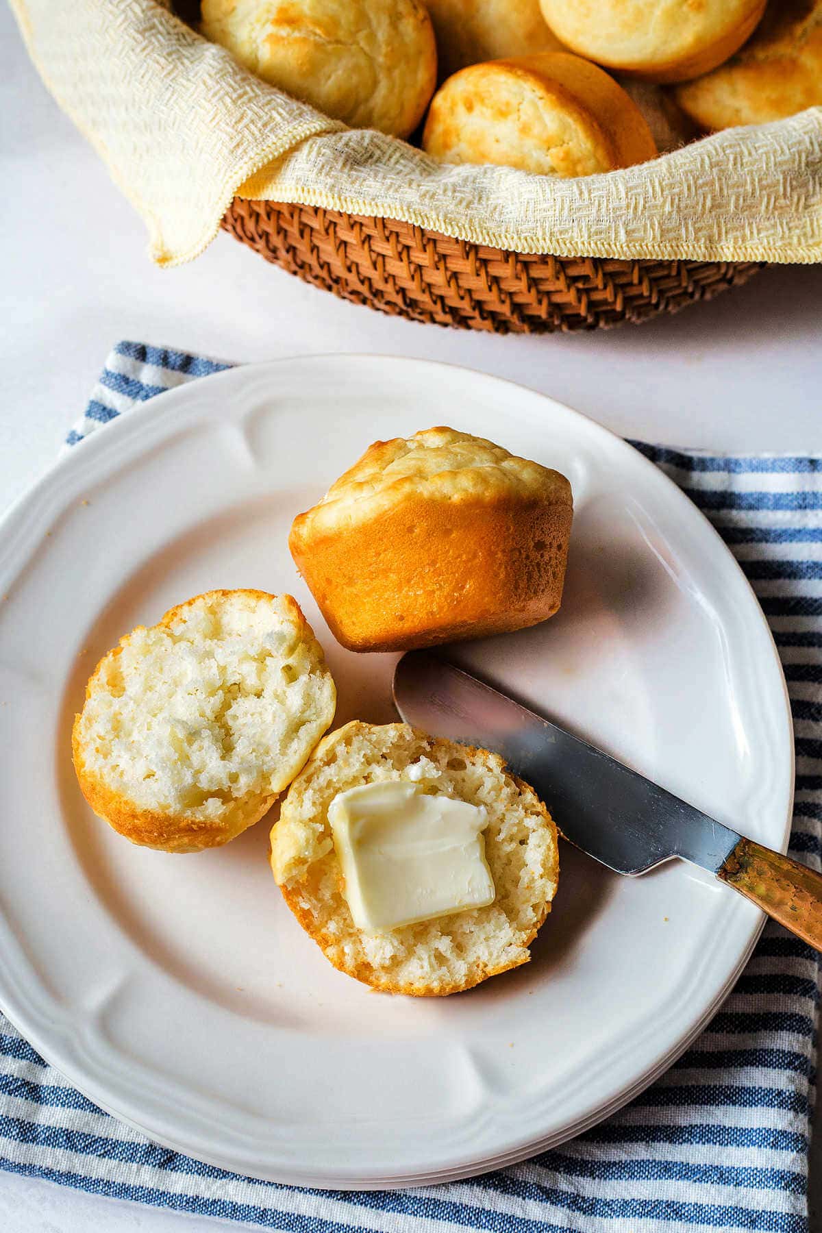 A split open mayo roll with a pat of butter on a plate with a basket of muffins in the background.