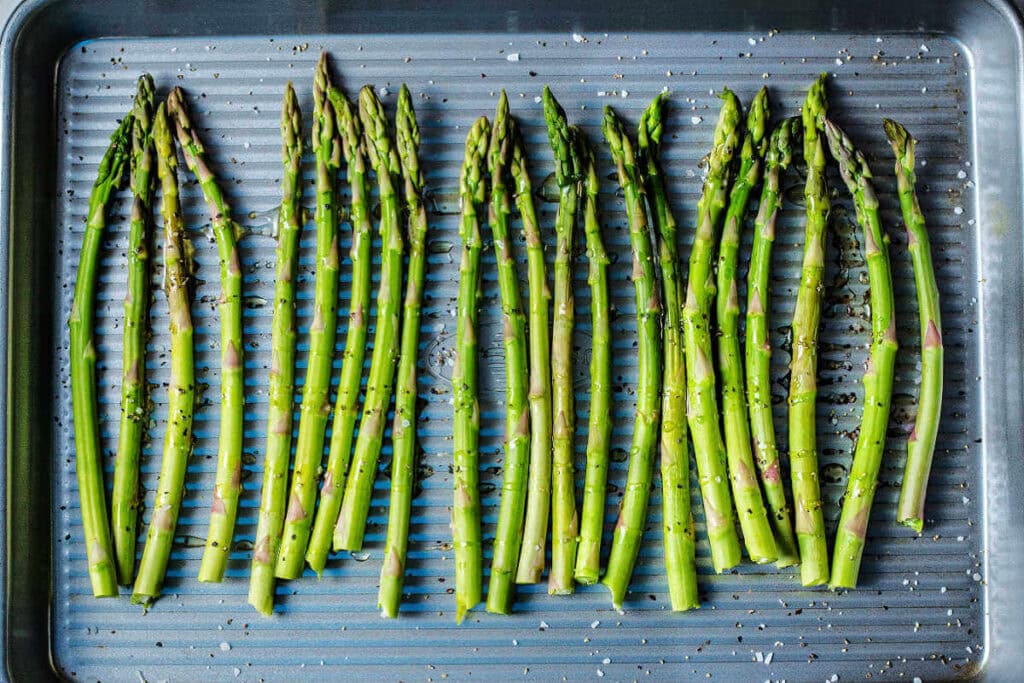 Pieces of asparagus lined up on a sheet pan, seasoned with salt and pepper.